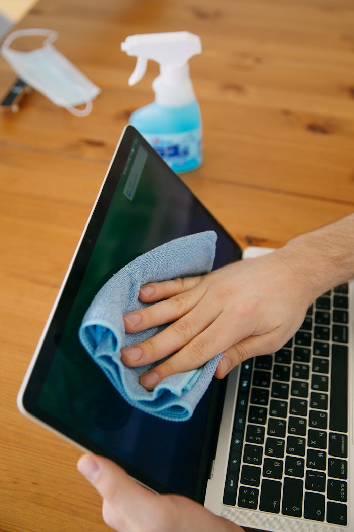 services-03 A person wipes a laptop screen using a blue cloth and spray bottle on a wooden table for hygiene.